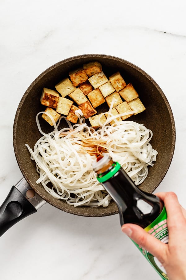 tou and rice noodles in a frying pan with a hand pouring soy sauce into the pan
