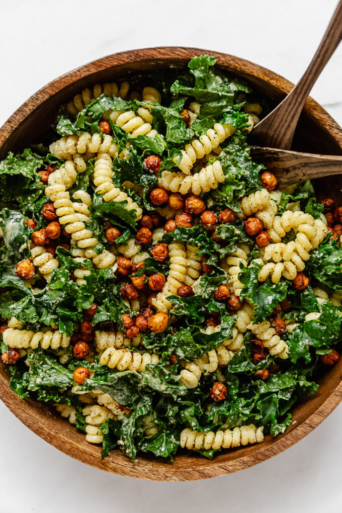 kale caesar pasta salad in a wood mixing bowl with 2 wood spoons